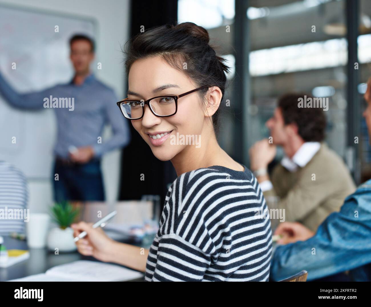 Dieser Job ist ein Starthilfe für meine Karriere. Porträt einer attraktiven jungen Frau, die in einem Büro mit Kollegen im Hintergrund sitzt. Stockfoto