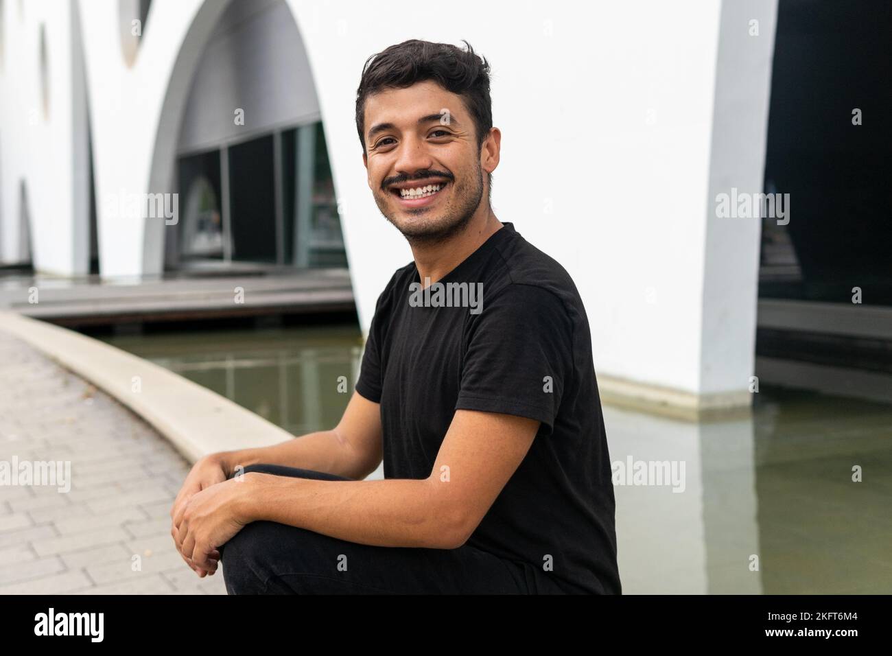 Ganzer Körper von positiven männlichen in schwarzen casual Outfit sitzt neben Brunnen Blick auf die Kamera Stockfoto