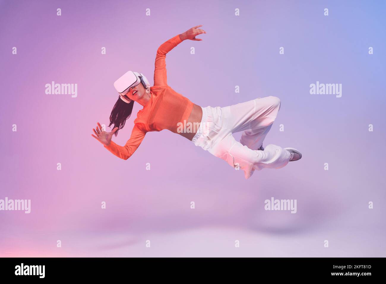 Ganzkörper-Weibchen im VR-Helm gestikuliert und fällt vor violettem Hintergrund, während sie den Cyberspace erkunden Stockfoto