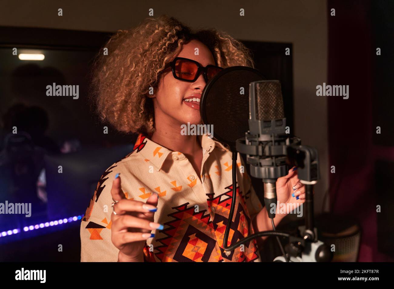 Junge Sängerin mit Afro-Frisur, die im professionellen Aufnahmestud gegen das strahlend orange Licht steht, während sie während der Probe Lied auf dem Mikrofon singt Stockfoto