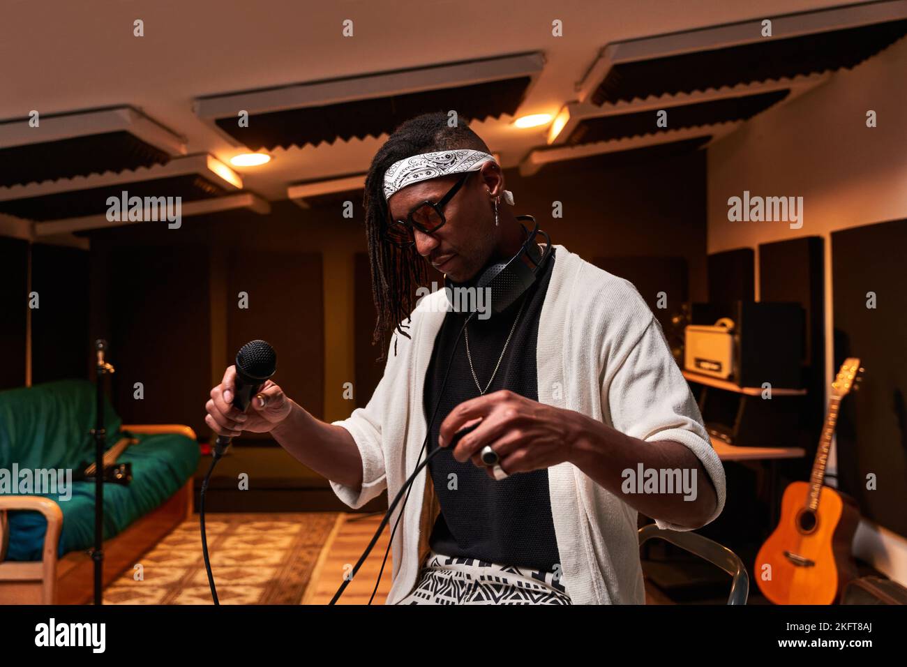 Afroamerikanischer Sänger in Bandana und Sonnenbrille, der im modernen Aufnahmestudio steht und den Mikrofondraht während der Probe justiert Stockfoto