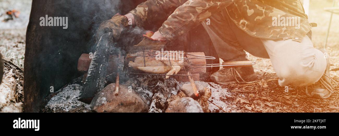 Mann Survivalist kocht Braten Huhn Fleisch Lebensmittel werden auf dem Grill auf schwelende Kohlen oder Emder vom Lagerfeuer auf dem Boden gebraten. Grill in Campingbedingungen Stockfoto