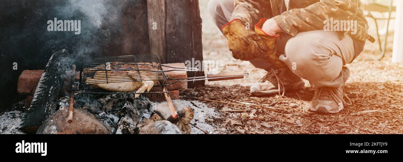 Mann Survivalist kocht Braten Huhn Fleisch Lebensmittel werden auf dem Grill auf schwelende Kohlen oder Emder vom Lagerfeuer auf dem Boden gebraten. Grill in Campingbedingungen Stockfoto