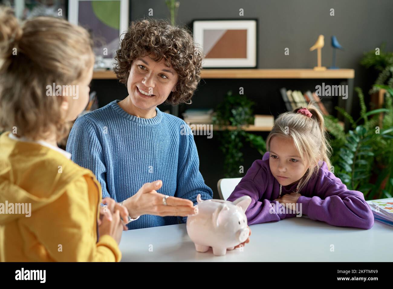 Mutter zeigte auf Sparschwein und lehrte ihre Kinder, Geld zu sparen, während sie am Tisch im Zimmer saßen Stockfoto