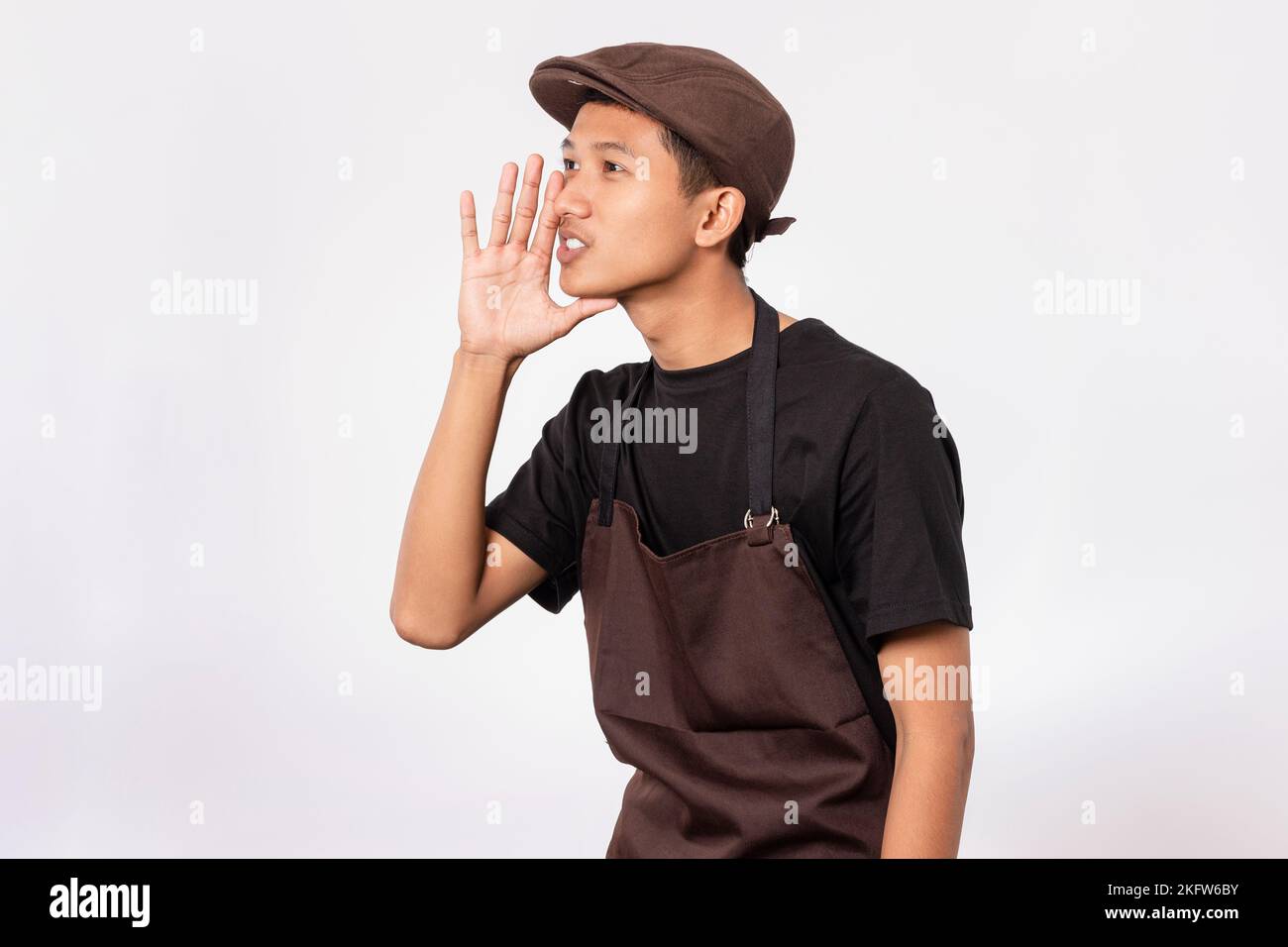 Ein hübscher, asiatischer Barista mit brauner Schürze und schwarzem T-Shirt, isoliert auf weißem Hintergrund, flüstert und erzählt das Geheimnis Stockfoto