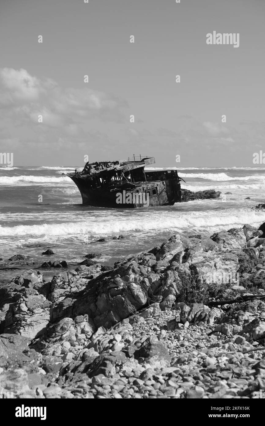 Eine vertikale Graustufe eines Schiffswracks in der Küstenlandschaft, die von einem Frachtschiff geschütteln wird Stockfoto