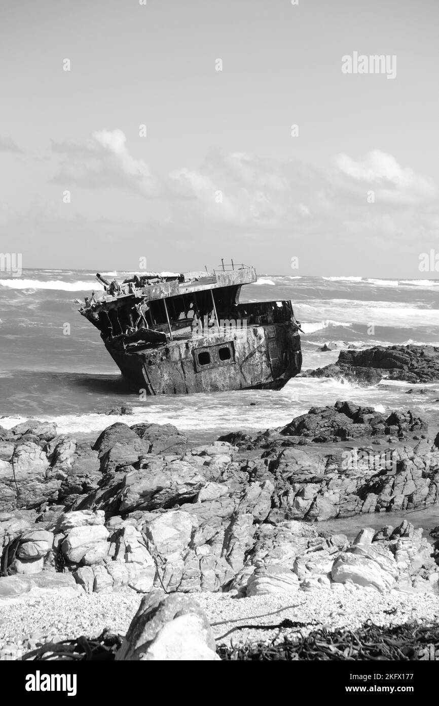 Eine vertikale Graustufe eines Schiffswracks in der Küstenlandschaft, die von einem Frachtschiff geschütteln wird Stockfoto