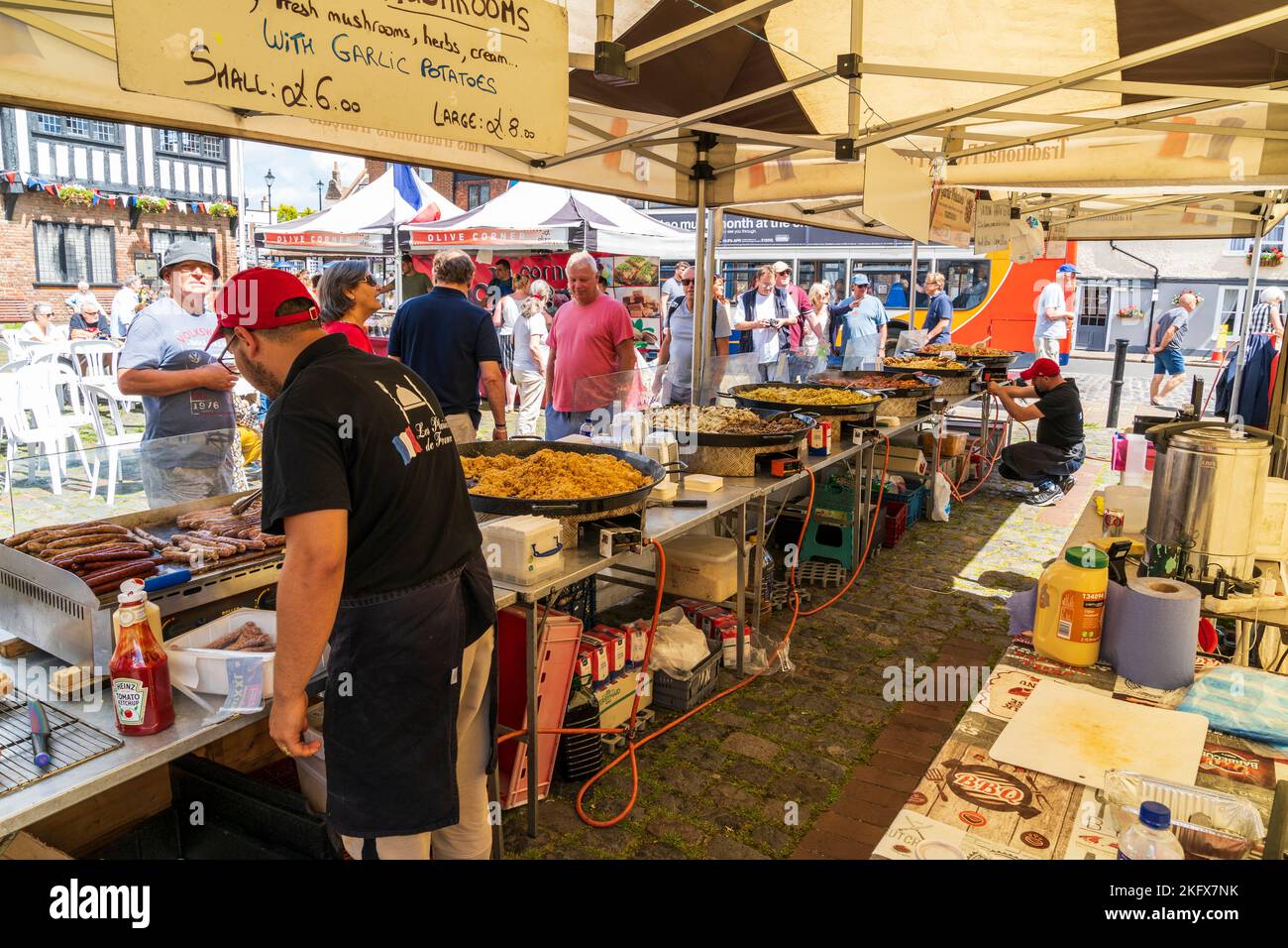 Blick von hinten auf einen offenen Imbissstand auf einem französischen Markt in England mit verschiedenen Pfanne Pfannen von Lebensmitteln, die mit Menschen gekocht werden, die sie betrachten. Stockfoto
