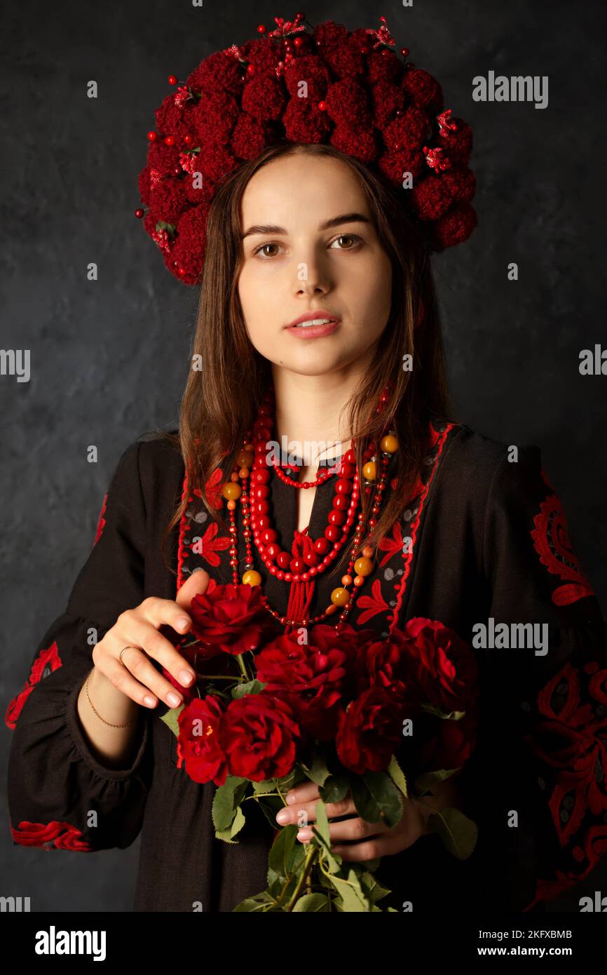Schöne ukrainische Frau in Kranz und Nationalkleid mit einem Strauß roter Rosen auf schwarzem Hintergrund Stockfoto