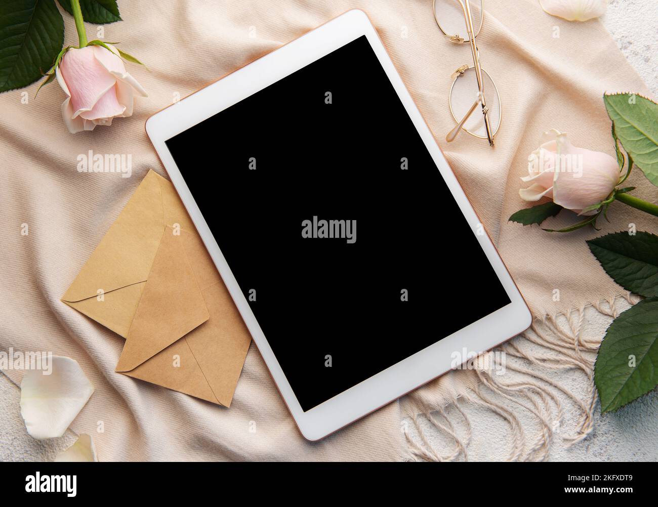 Minimalistischer Arbeitsplatz mit digitalem Tablet leerem Bildschirm-Mockup, gemütlichem Schal, Rosen-Blumen auf betontem Hintergrund. Draufsicht, flach liegend Stockfoto