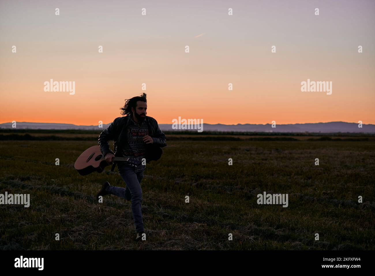 kaukasischer junger Mann mit langen Haaren in Pelzjacke, der bei Sonnenuntergang mit einer Gitarre durch das große einsame Feld läuft Stockfoto