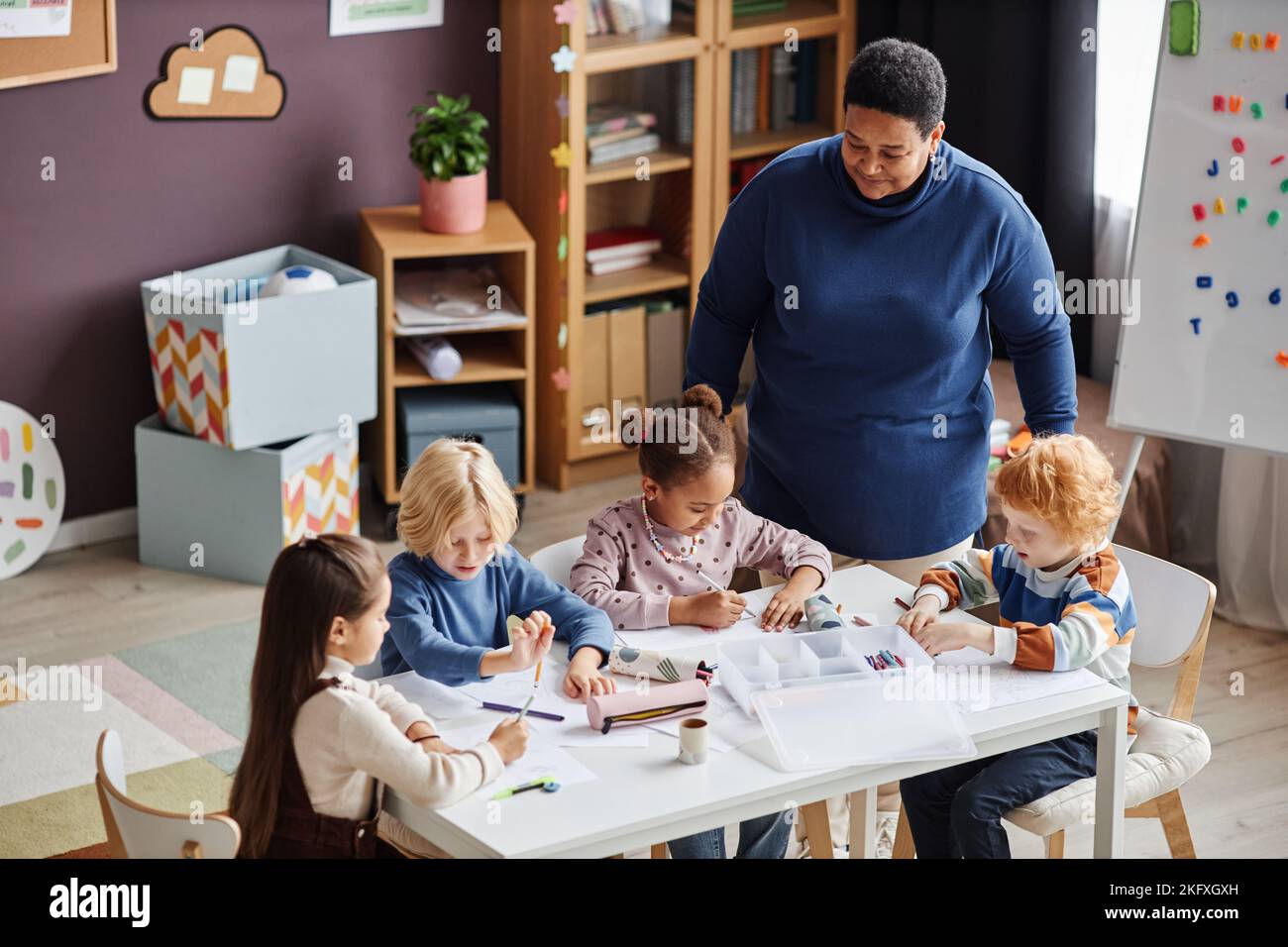 Gruppe von liebenswerten Lernenden der Kindergarten sitzen am Tisch bei der Lektion, während reife afroamerikanische Lehrerin neben ihnen stehen Stockfoto