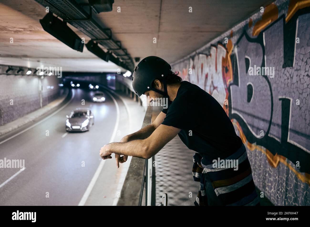 Der junge kaukasische Mann steht mit Helm auf dem Kopf und lehnt sich auf das Geländer eines Gehwegs in einem Tunnel mit viel Verkehr in der Stadt Stockfoto