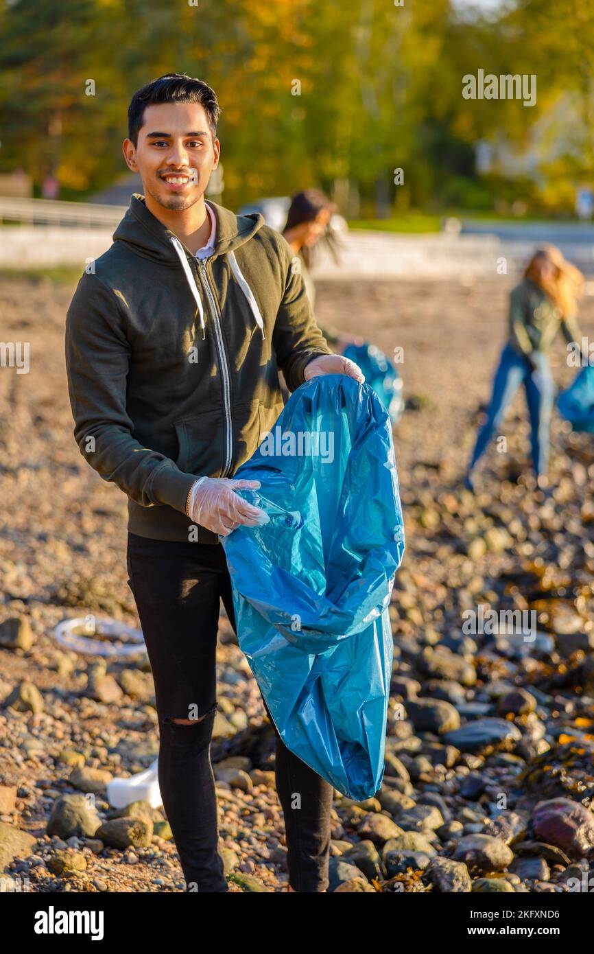 Der junge Mann sammelt am sonnigen Tag mit einer Gruppe von Freiwilligen Müll in der Tasche Stockfoto