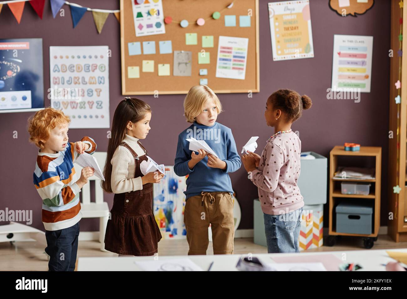 Gruppe von vier interkulturellen kleinen Lernenden des Kindergartens in Casualwear, die Papierflugzeuge spielen und in der Pause im Klassenzimmer interagieren Stockfoto