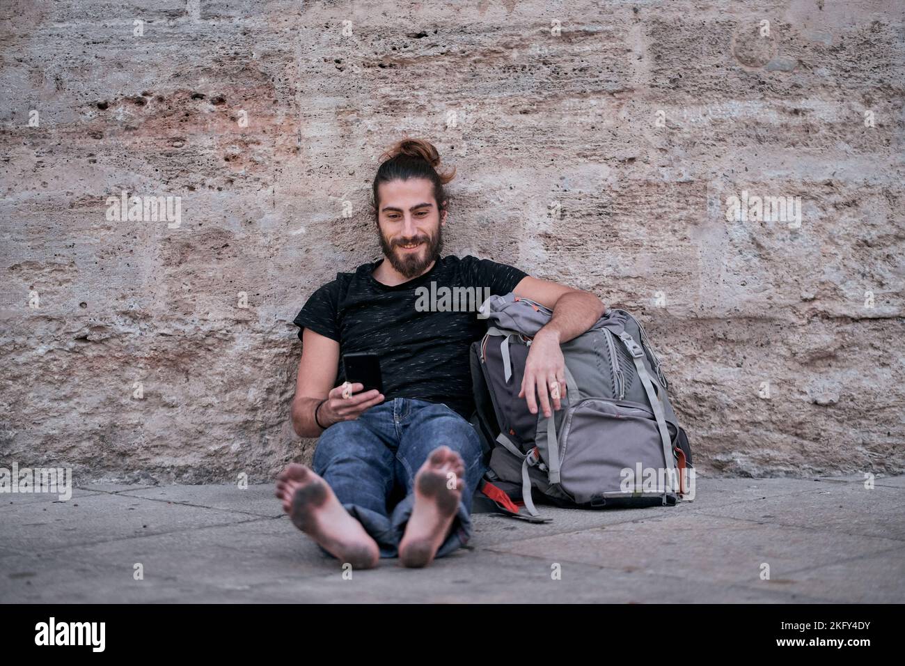 kaukasischer Kerl mit langen Haaren Beine gestreckt barfuß Blick auf die Kamera mit seinem Smartphone in der Hand sitzend auf seinem großen Rucksack auf der gelehnt Stockfoto