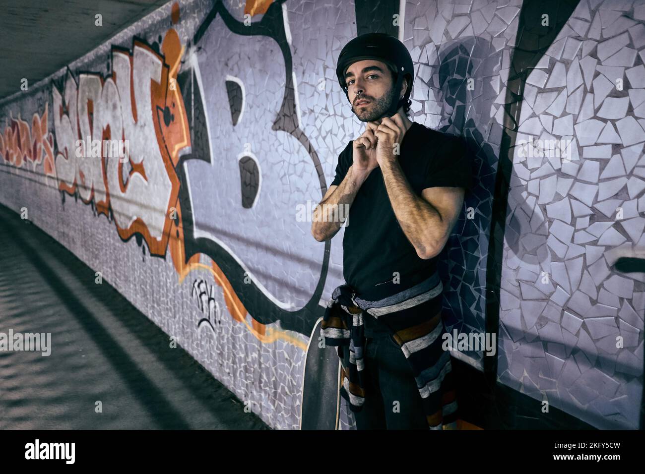 Der junge kaukasische Mann steht und bindet den Helm, den er auf seinem Kopf trägt und lehnt sich an die Wand eines Tunnels und blickt auf die Kamera neben einem Skateboard Stockfoto