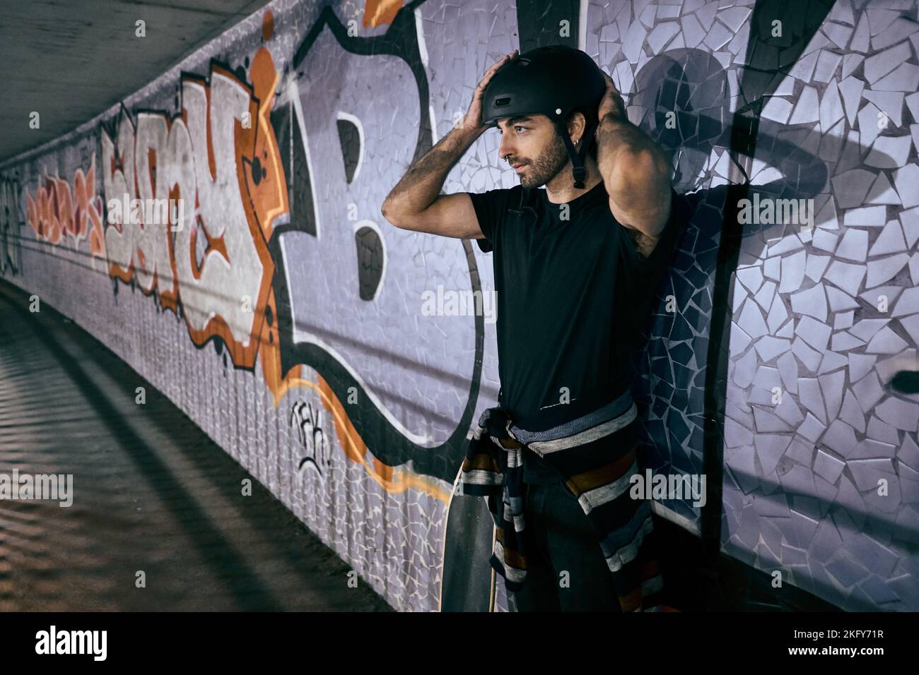Der junge kaukasische Mann steht und setzt einen Helm auf den Kopf, der sich an einer Tunnelwand neben einem Skateboard in der Stadt lehnt Stockfoto