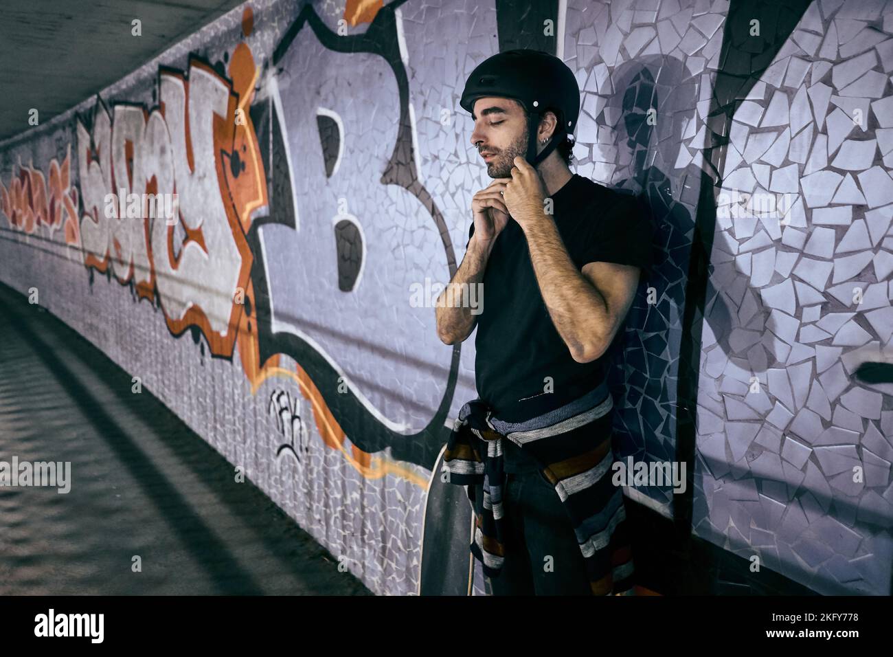 Der junge kaukasische Mann steht und bindet den Helm, den er auf seinem Kopf trägt, an der Wand eines Tunnels neben einem Skateboard in der Stadt gelehnt Stockfoto