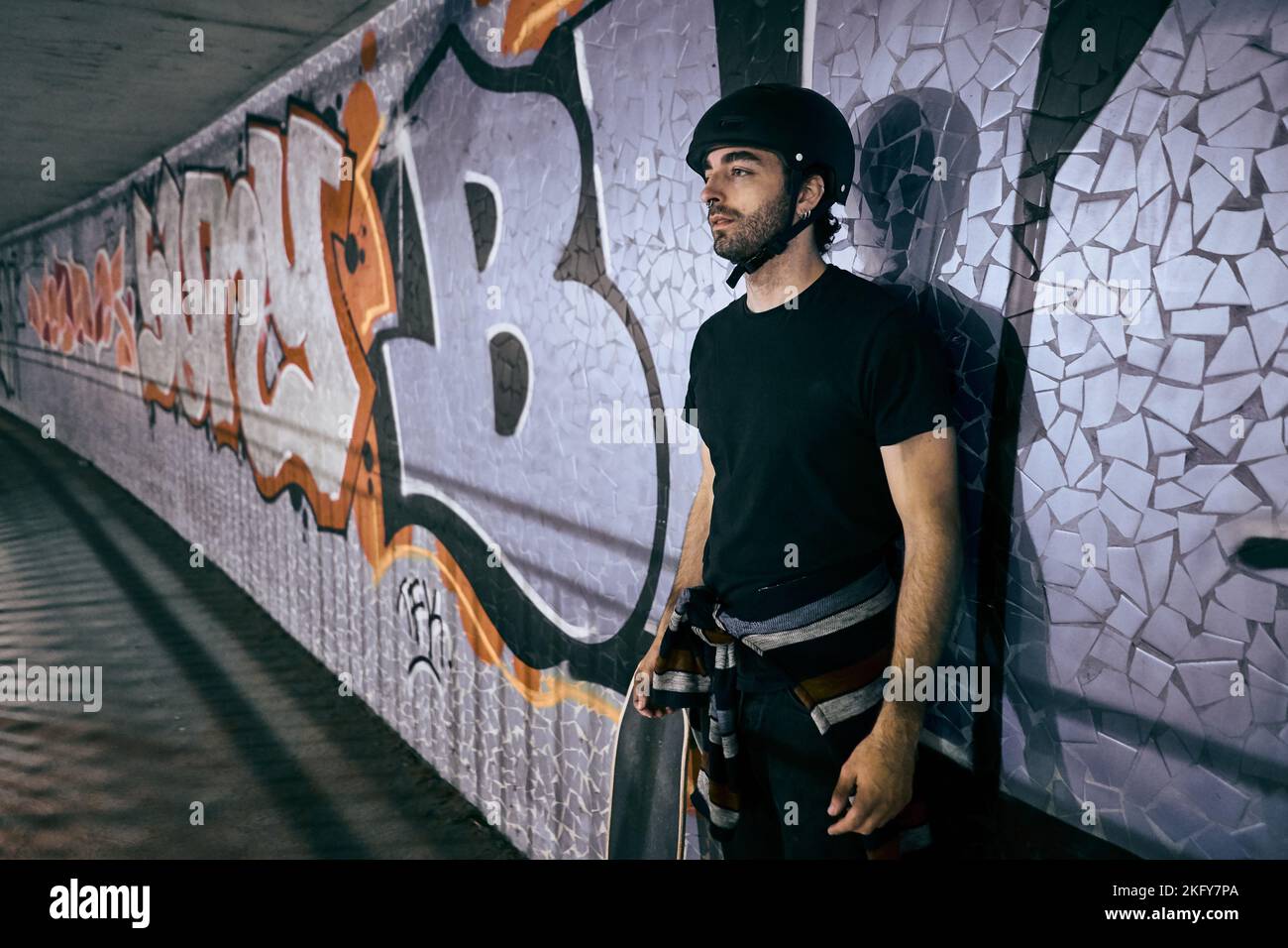 Der junge Kaukasusmann steht mit Helm auf dem Kopf und lehnt sich an die Wand eines Tunnels neben einem Skateboard in der Stadt Stockfoto