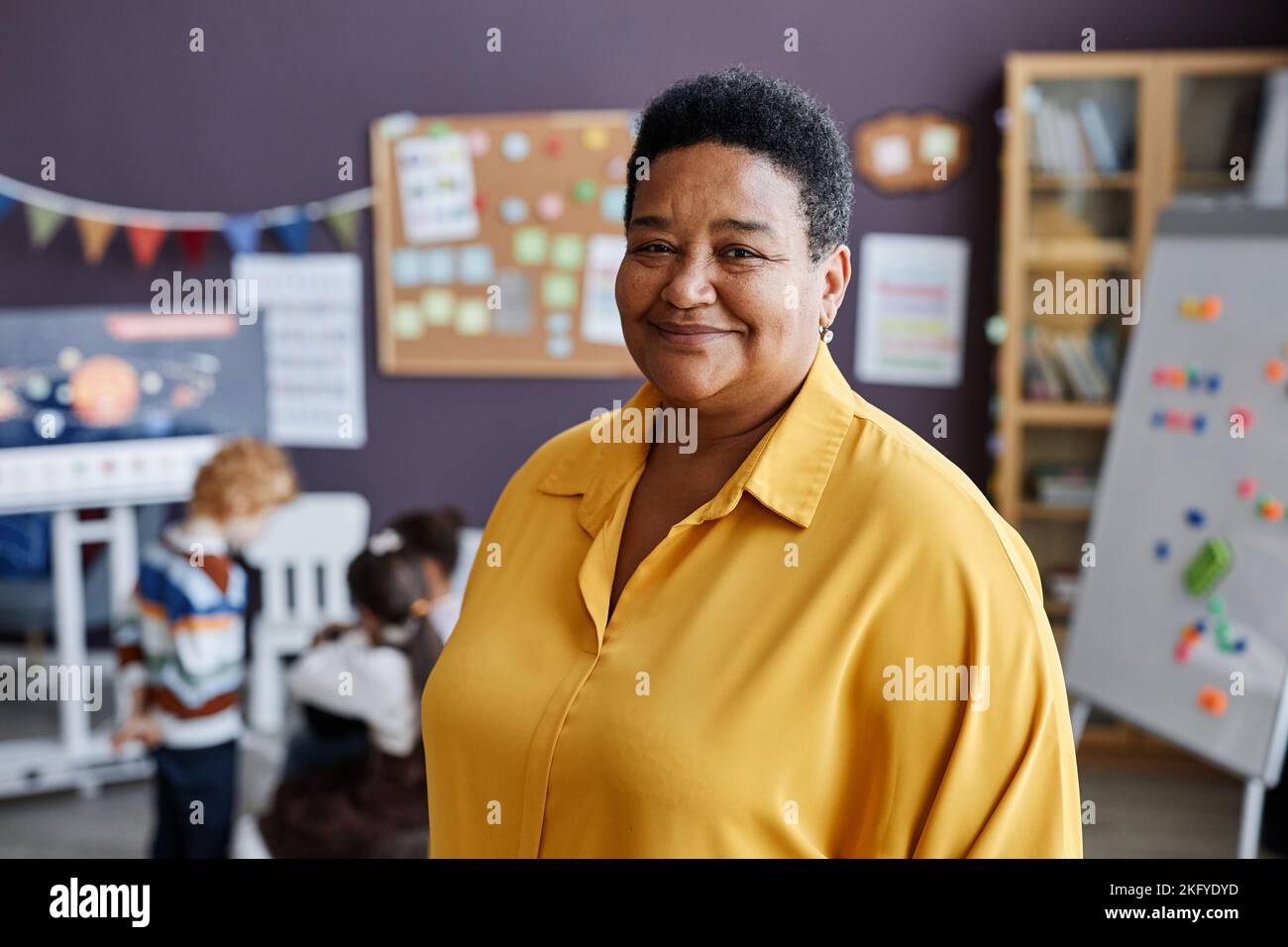 Erfolgreiche professionelle reife Lehrerin der Kindergartenschule, die Kamera anschaut, während sie gegen eine Gruppe von kleinen Lernenden steht Stockfoto