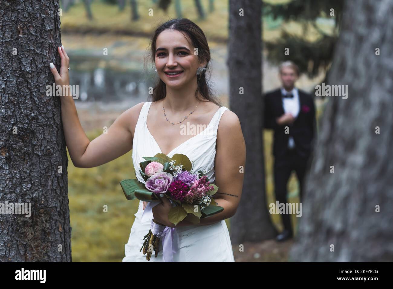 Fotoshooting mit der Herbstzeit. Mittelgroße Außenaufnahme einer atemberaubenden türkischen Frau Mitte 20s, die einen Baum berührte, lächelte und ihren Brautstrauß hielt. Verschwommene Silhouette des eleganten skandinavischen Bräutigams im Hintergrund. Hochwertige Fotos Stockfoto