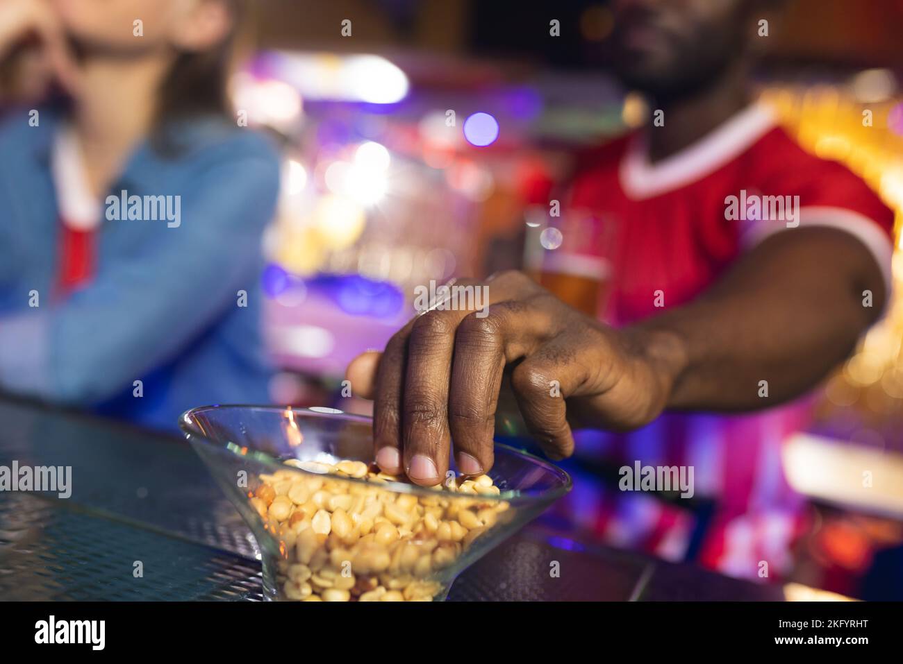 Zwei verschiedene männliche und weibliche Freunde an der Bar, Mann schnappt sich Erdnüsse aus der Schüssel, selektiver Fokus Stockfoto