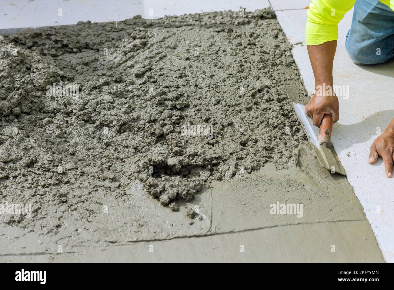 Der Arbeiter hält eine Stahlkelle, während er den neu gegossenen Betonweg im Baubereich glättet und ebnet Stockfoto