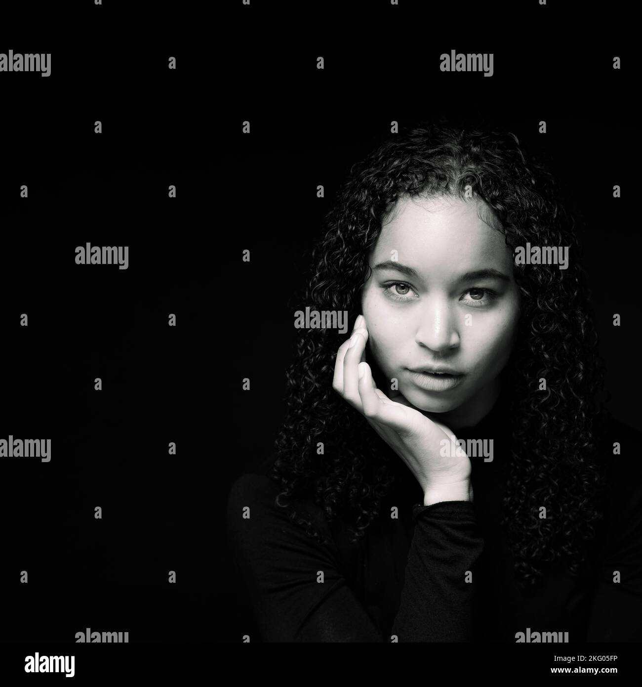 Nahaufnahme der jungen multirassischen Frau mit der Hand auf Chin | Schwarzer Hintergrund mit Copy Space auf der linken Seite Stockfoto