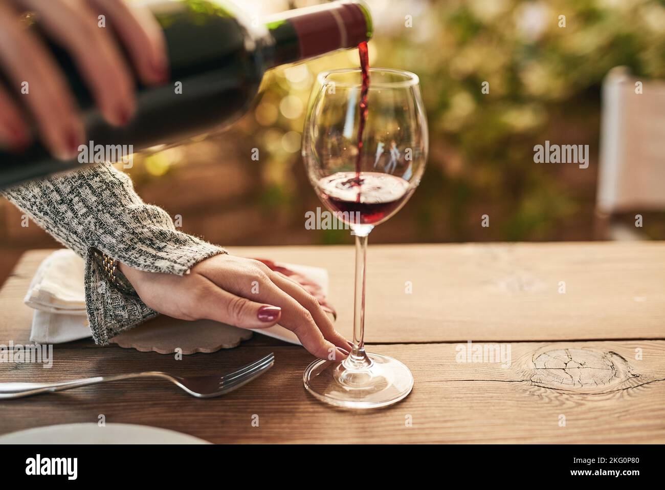 Glas, Flasche und Hand einer Frau, die Wein an einen Tisch gießt, ruhig und zufrieden, während sie ihre Freizeit auf einer Terrasse genießen. Rotwein, Hände und Dame entspannen sich mit Stockfoto