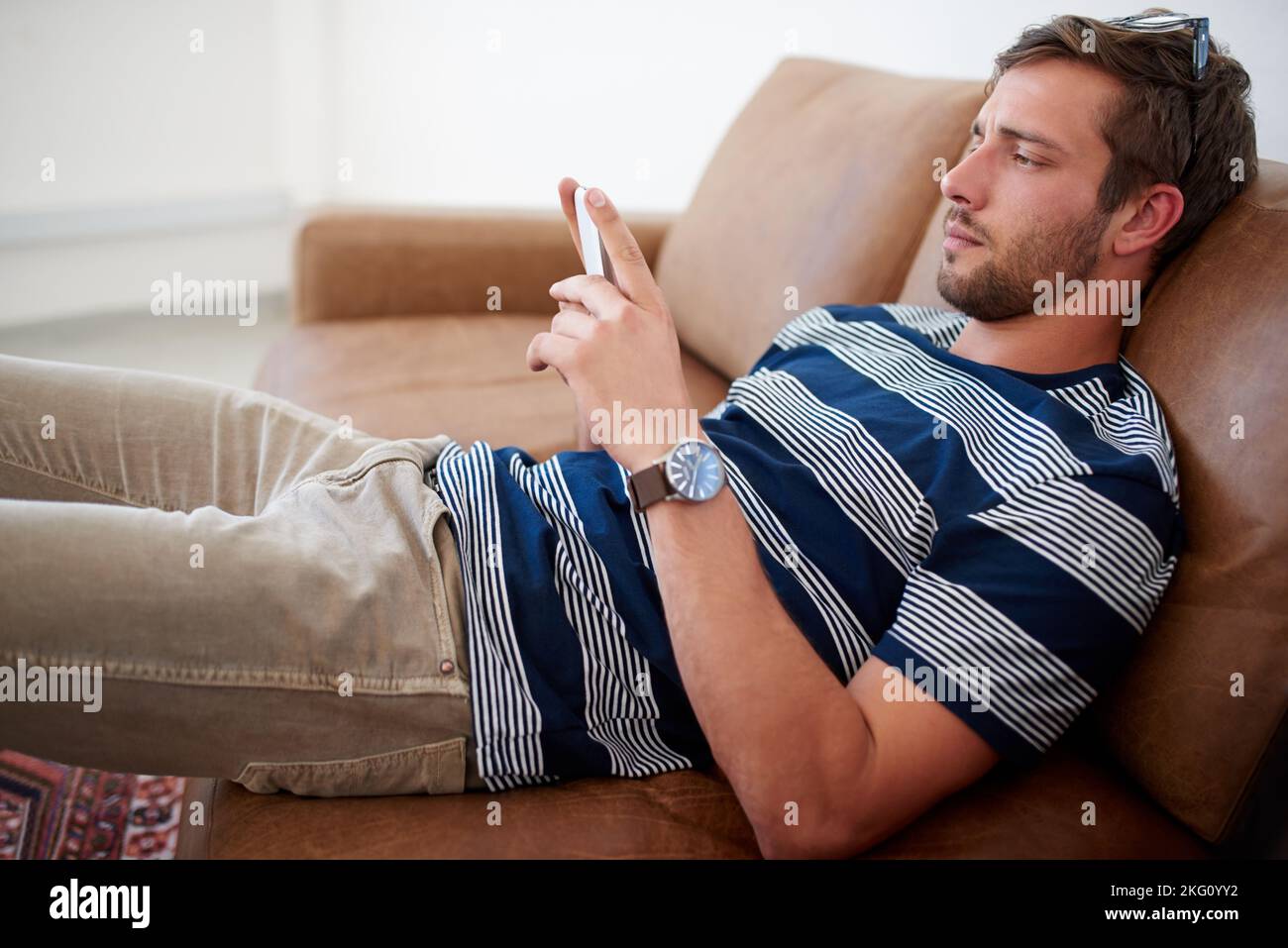 Geselligkeit in seiner Pause. Hübscher junger Mann, der auf einer Couch faulenzt, während er auf seinem Telefon SMS schreibt. Stockfoto