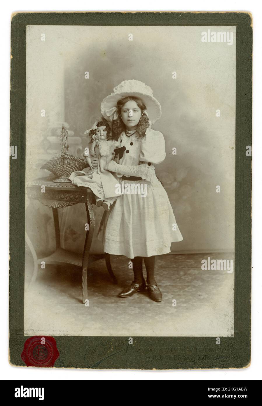 Edwardian Ära Cabinet Card Studio Porträt eines verwöhnten privilegierten, wohlhabenden edwardianischen Mädchens der mittleren oder oberen Klasse, das eine Puppe hält, die ein porzellangesicht hat, vielleicht ein neues Geburtstagsgeschenk oder einen wertvollen Besitz hat oder eine Studio-Requisite sein könnte. Auf der Rückseite steht Clara R. J. Whant, 11 1909 Jahre alt. Das Mädchen trägt teure Kleidung - einen großen Hut mit breiter Krempe, wie es damals Mode war, und einen weißen Rock mit Puffärmeln. Sie hat lange Haare gekräuselt, die in Bögen gebunden sind. Aus dem Studio von Edward Sharp in Islington und Westminster Bridge London. Großbritannien vom 1909. Stockfoto