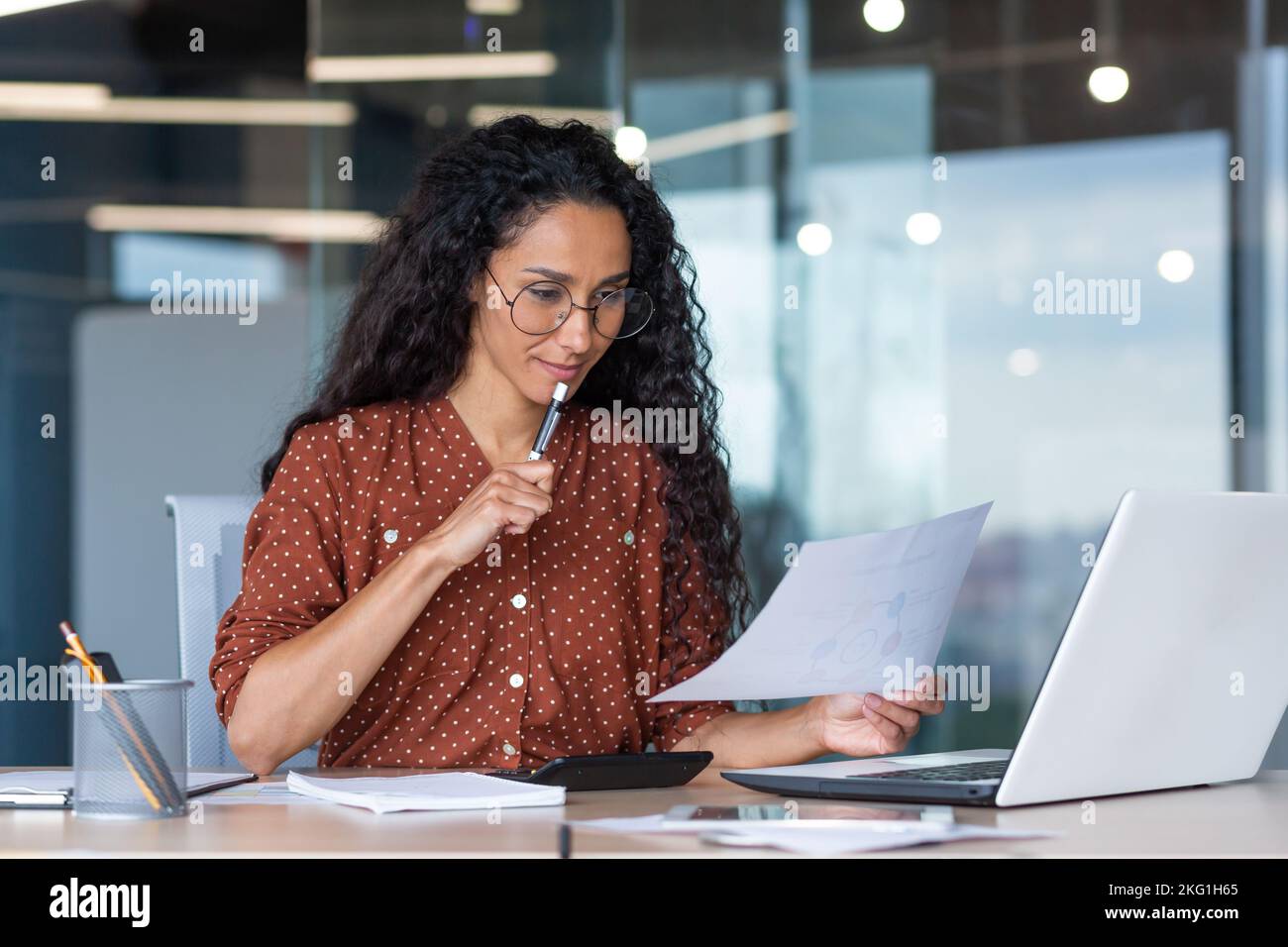 Schöne hispanische Frau im Büro, die Papierkram macht, erfolgreiche Geschäftsfrau Finanzierin, die im Büro am Tisch mit Laptop sitzt, bei der Arbeit mit Verträgen und Rechnungen, über Entscheidungen nachdenkt. Stockfoto