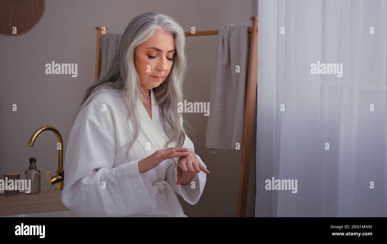 Schöne Oma 60s Dame alt Senior grauhaarige weibliche kaukasische Frau im Schlafzimmer reiben faltige Hände Anwendung natürliche Handcreme feuchtigkeitsspendend Stockfoto