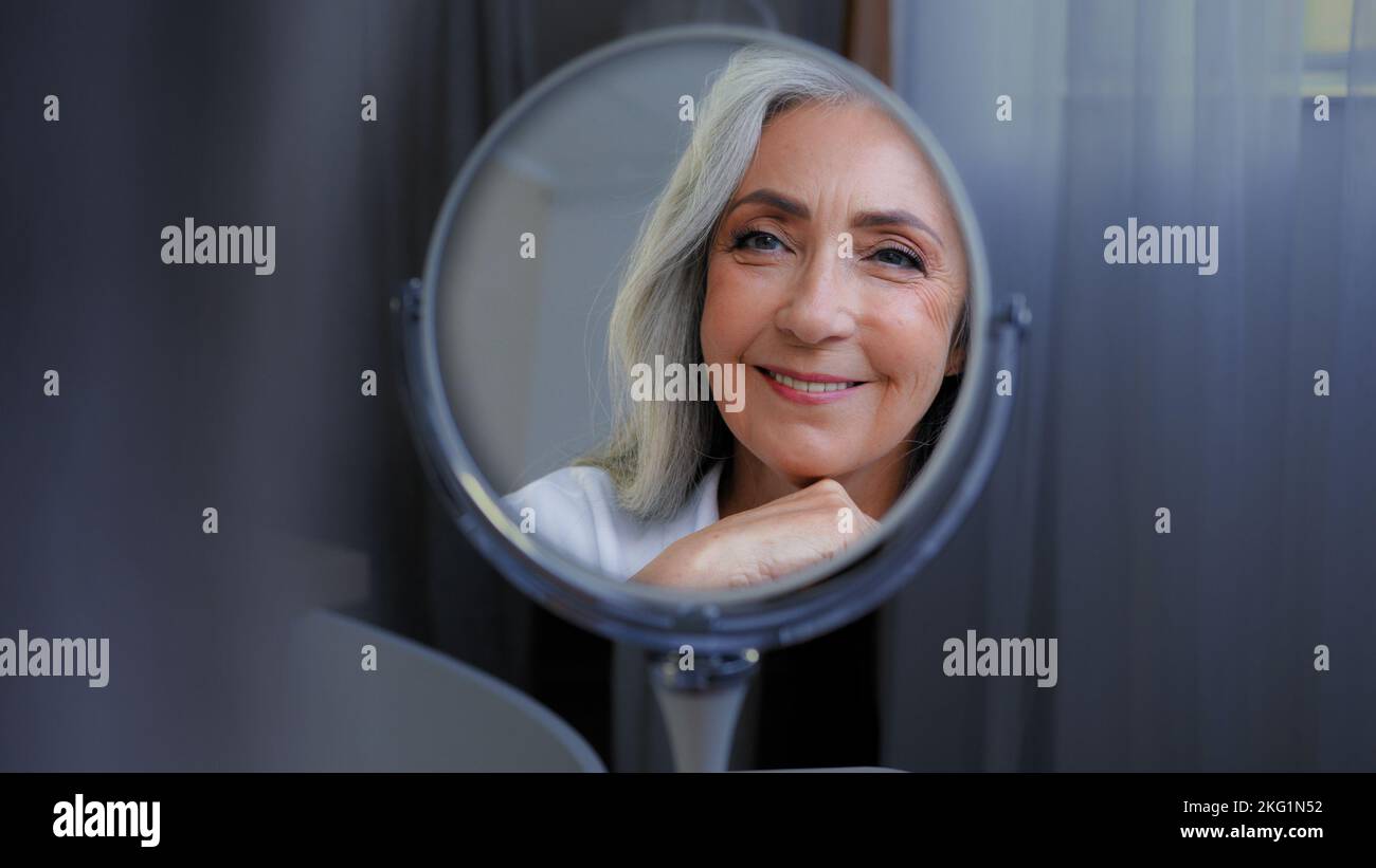 Lächelnd schönes feminines weibliches Gesicht 50s ältere Frau im mittleren Alter genießen Gesichtshaut Blick auf Spiegelreflexion Verwöhnung. Alte alternde trockene Hautpflege Stockfoto