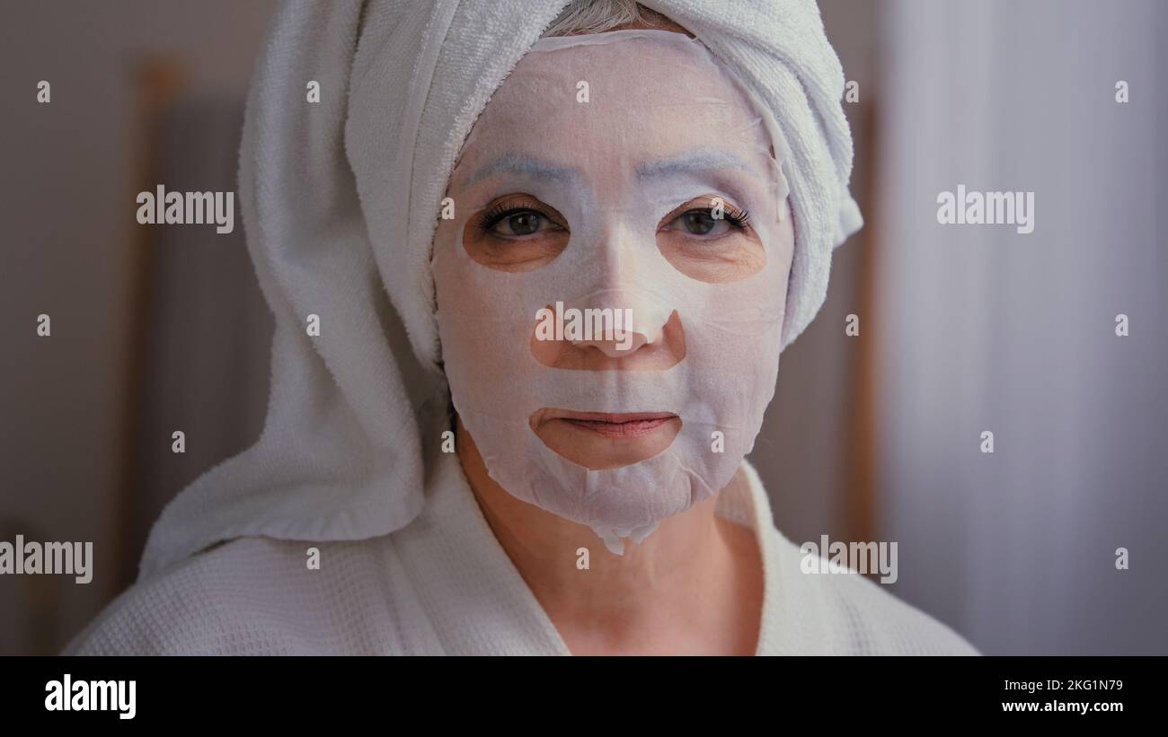Alte ältere ältere kaukasische Frau gelten Hautpflege textile Hautmaske auf weibliches Gesicht zeigt ok gute Schönheit Kosmetologie Verfahren im Badezimmer 60s Dame Stockfoto