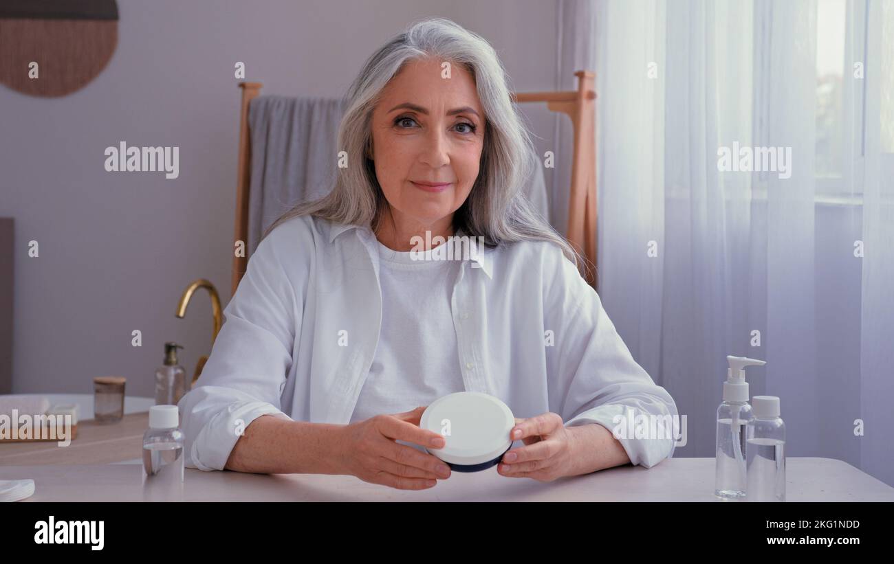 Kaukasische alte ältere ältere ältere 50s Frau mit grauem Haar vlogger in Innenräumen zu Hause sprechen Blick auf die Kamera zeigen Beauty-Produkte Creme Stockfoto