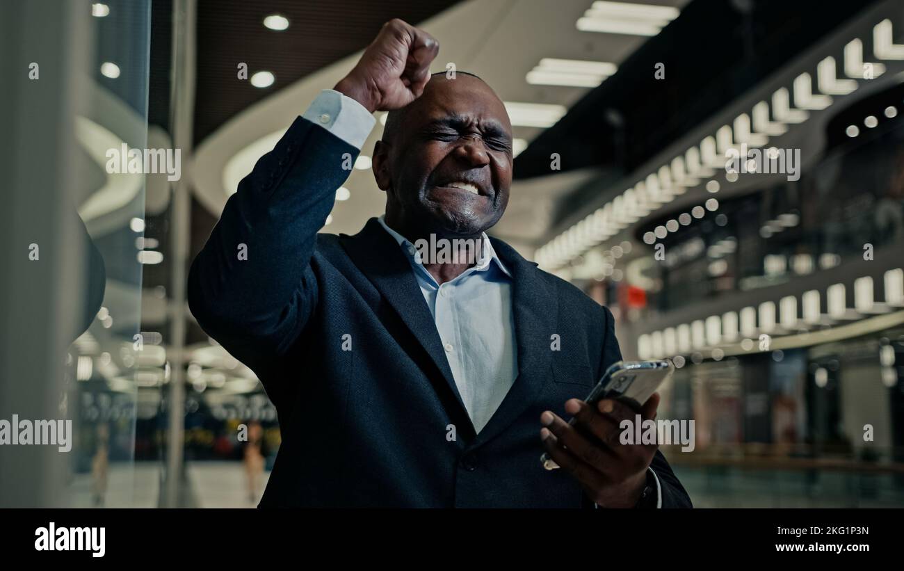 Glücklicher Sieger Erfolg Erwachsener Geschäftsmann afrikanischer Mann mittleren Alters Angestellter Unternehmer drinnen im Büro Einkaufszentrum Firma gewinnen mit Handy Sieg Stockfoto