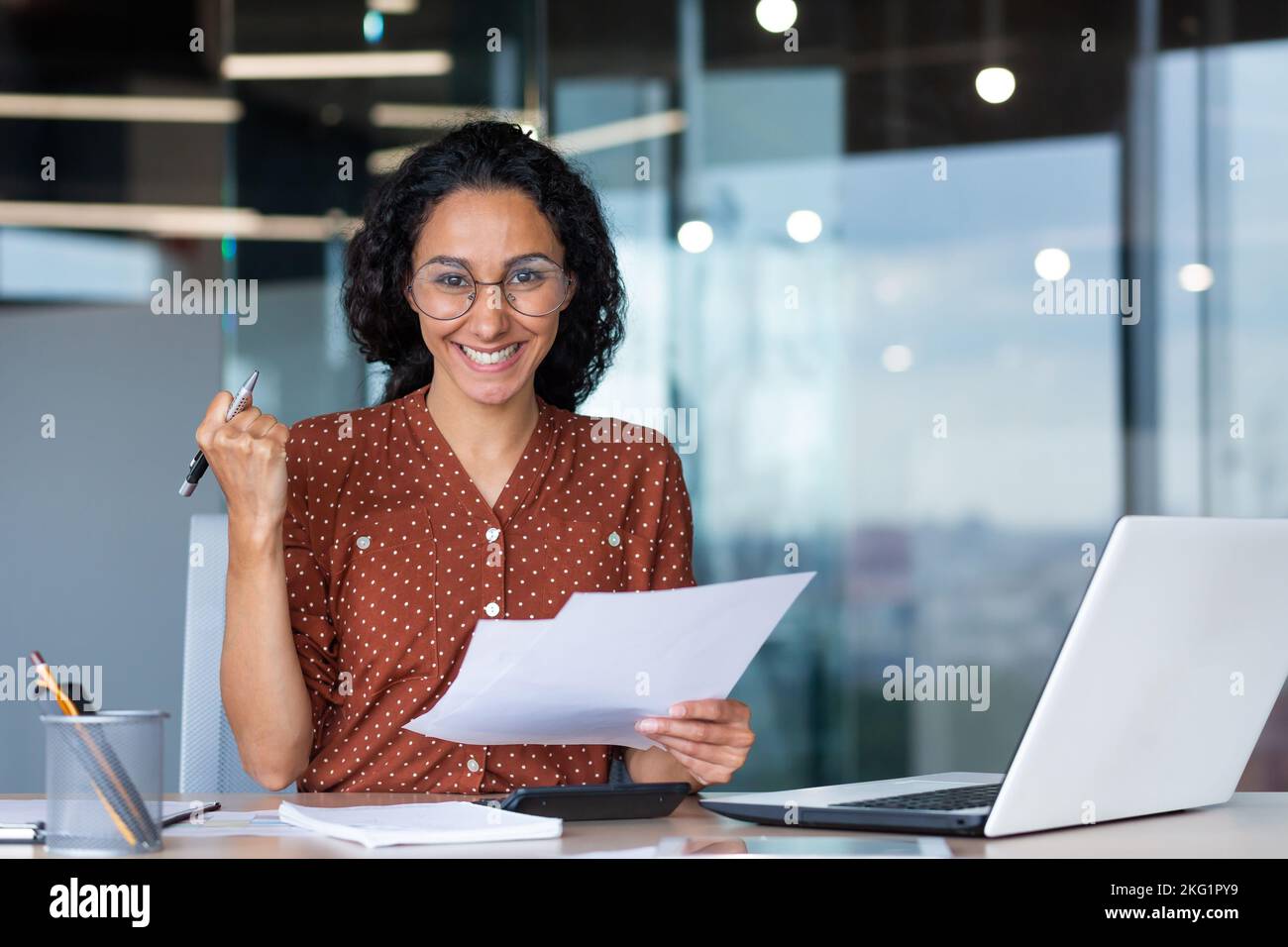 Erfolgreiche und glückliche Geschäftsfrau zufrieden mit den Erfolgen, lächelnd und mit Blick auf die Kamera, die Dokumente und Finanzberichte hält, hispanic-Frau, die im Büro am Arbeitsplatz mit einem Laptop arbeitet. Stockfoto