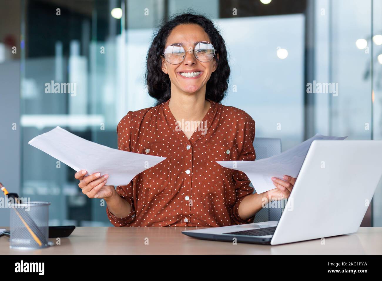 Erfolgreiche und glückliche Geschäftsfrau zufrieden mit den Erfolgen, lächelnd und mit Blick auf die Kamera, die Dokumente und Finanzberichte hält, hispanic-Frau, die im Büro am Arbeitsplatz mit einem Laptop arbeitet. Stockfoto