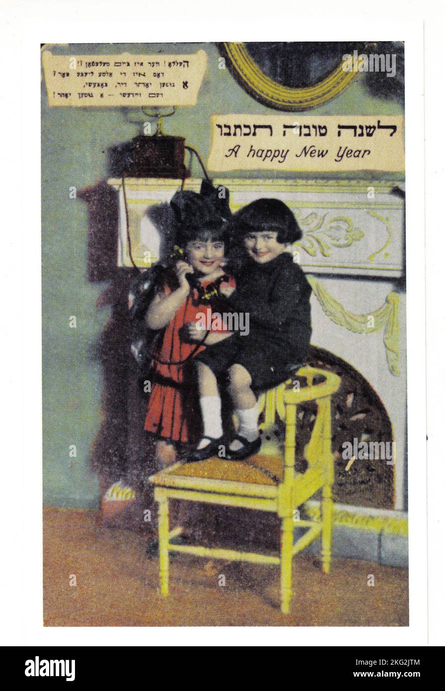 Eine jüdische Neujahrskarte aus dem frühen 20.. Jahrhundert, die hauptsächlich auf Jiddisch, aber auch mit hebräischer und englischer Schrift verfasst wurde. Es zeigt zwei kleine Kinder, die ihre Grüße auf einem altmodischen Telefon ausdehnen, eine relativ neue Erfindung für das Haus. Stockfoto