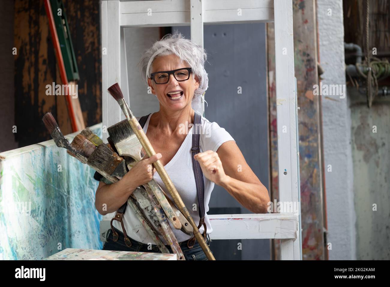 Ältere grauhaarige reife glückliche Künstlerin Frau mit Brille und großen Pinsel machen Faust Macht Geste, kopieren Raum Stockfoto
