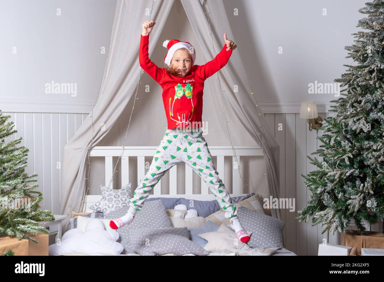 Happy lustige Baby Mädchen in Pyjamas und Santa Hut springt im Bett am Weihnachtsmorgen. Die Kinder genießen die Weihnachtsfeiertage. Das Kind hat Spaß Stockfoto