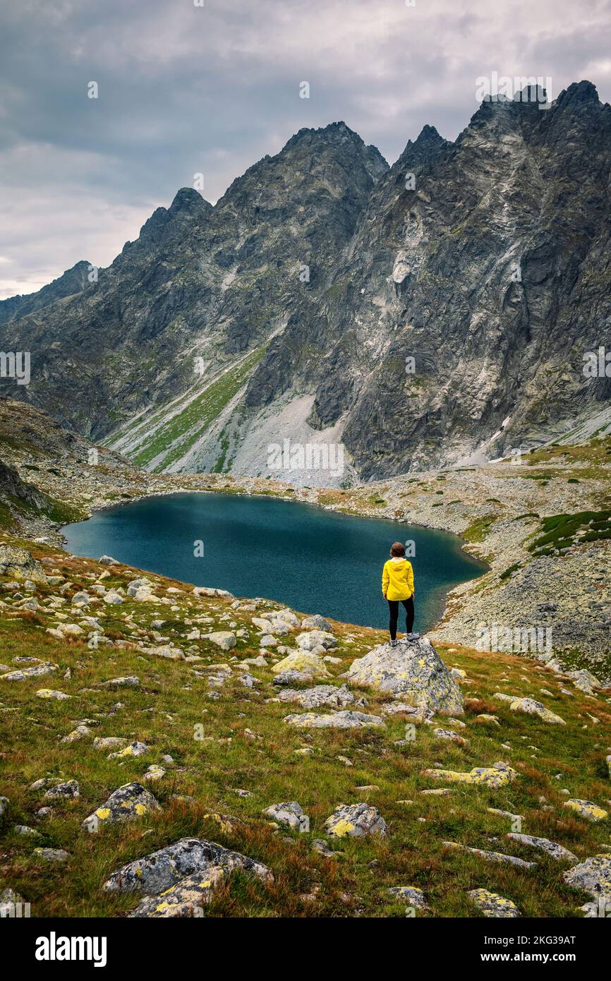 Wanderer Frau erkunden Sie die fantastische Landschaft und genießen Sie die Aussicht in der Nähe eines Bergsees in gelb in Jacke, hohe Tatra, Slowakei Stockfoto