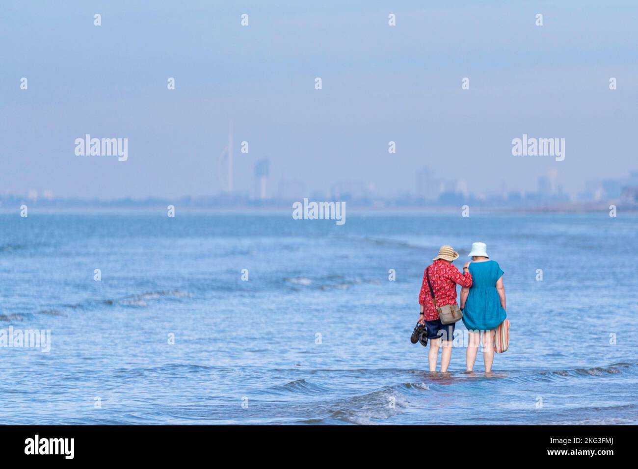 Zwei Damen paddeln im herrlichen Wasser des West Wittering Beach mit Blick auf Portsmouth's Spinnaker Tower. Stockfoto