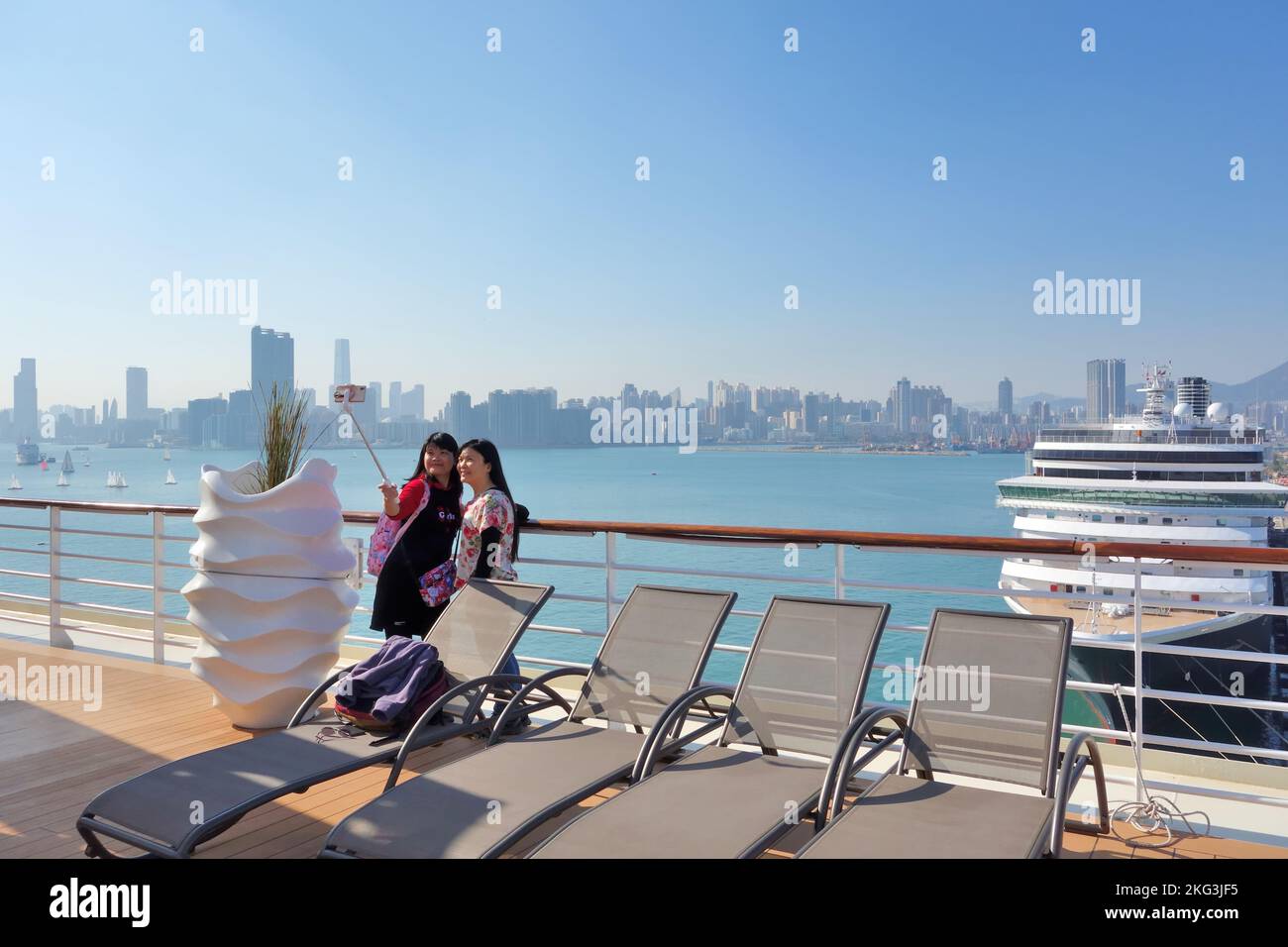 Zwei asiatische Mädchen verwenden einen Selfie-Stick und ein Mobiltelefon, um ein Urlaubsfoto auf einem Kreuzschiff zu machen, während sie in Hongkong, China, angedockt sind. Stockfoto