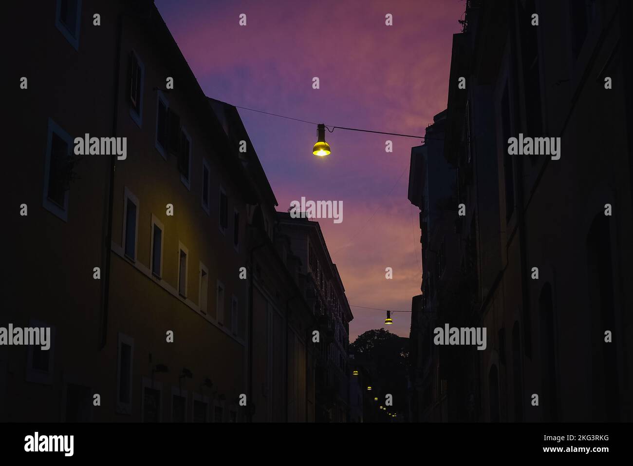 Dunkle und launische Straße in Rom, Italien, in der Dämmerung. Die Lampe leuchtet vor dem violett-pinken Abendhimmel. Gebäude im Schatten. Nostalgische und dramatische Postkarte. Stockfoto