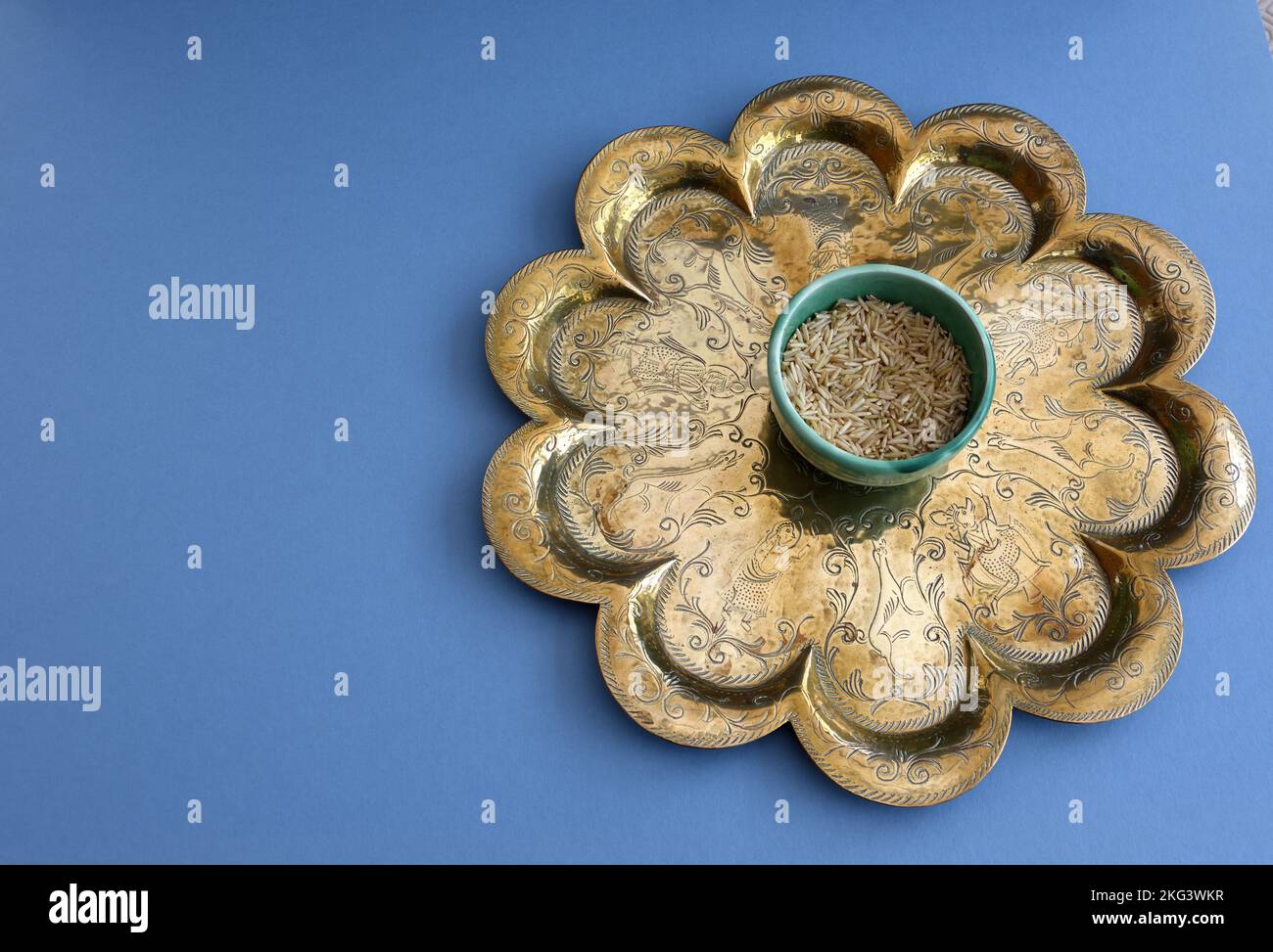 Basmati-brauner ungekochter Reis in grüner Keramik-Brannam-Schüssel auf einem antiken indischen Messingtablett auf blauem Hintergrund. Abgewogen zur Vorbereitung auf das Kochen a c Stockfoto