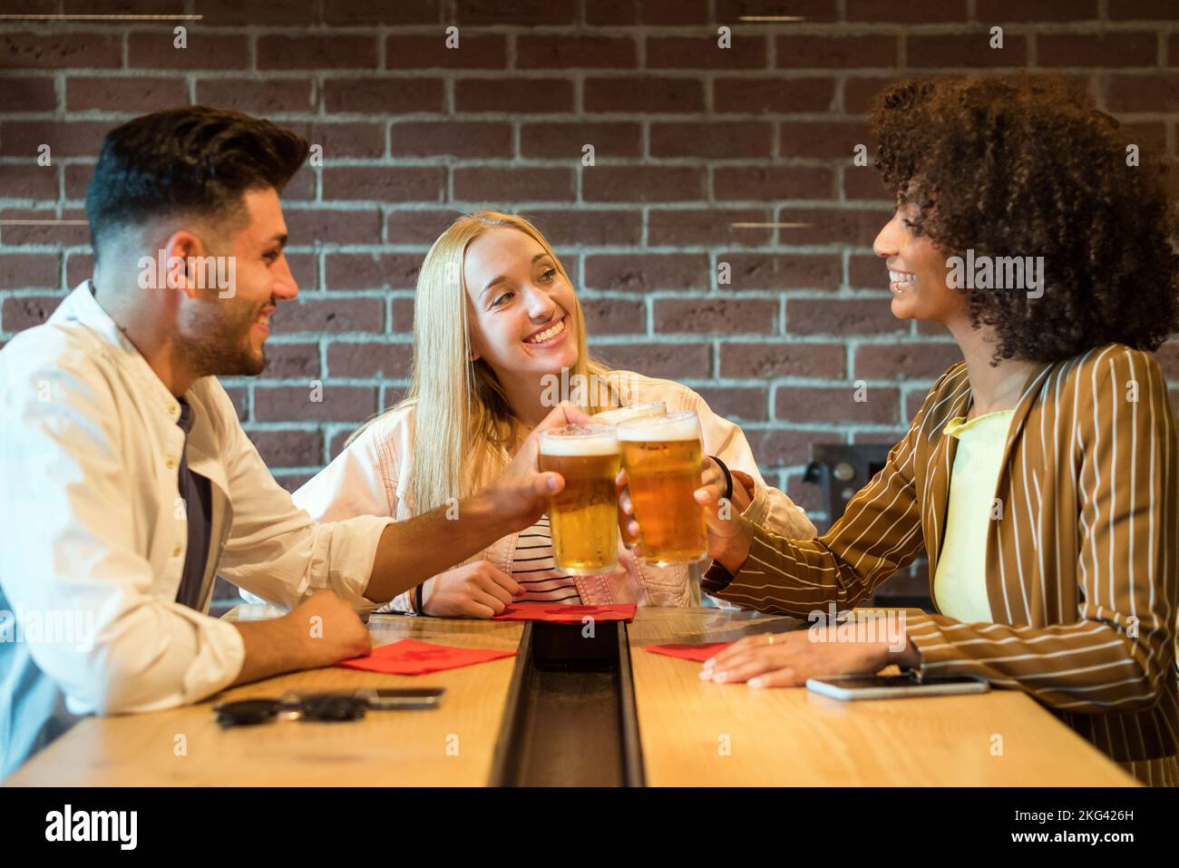 Lächelndes, multirassisches Jubeln mit Gläsern kalten, schäumenden Bieres, während man am Tisch in einem Pub in der Nähe einer Ziegelsteinmauer sitzt Stockfoto