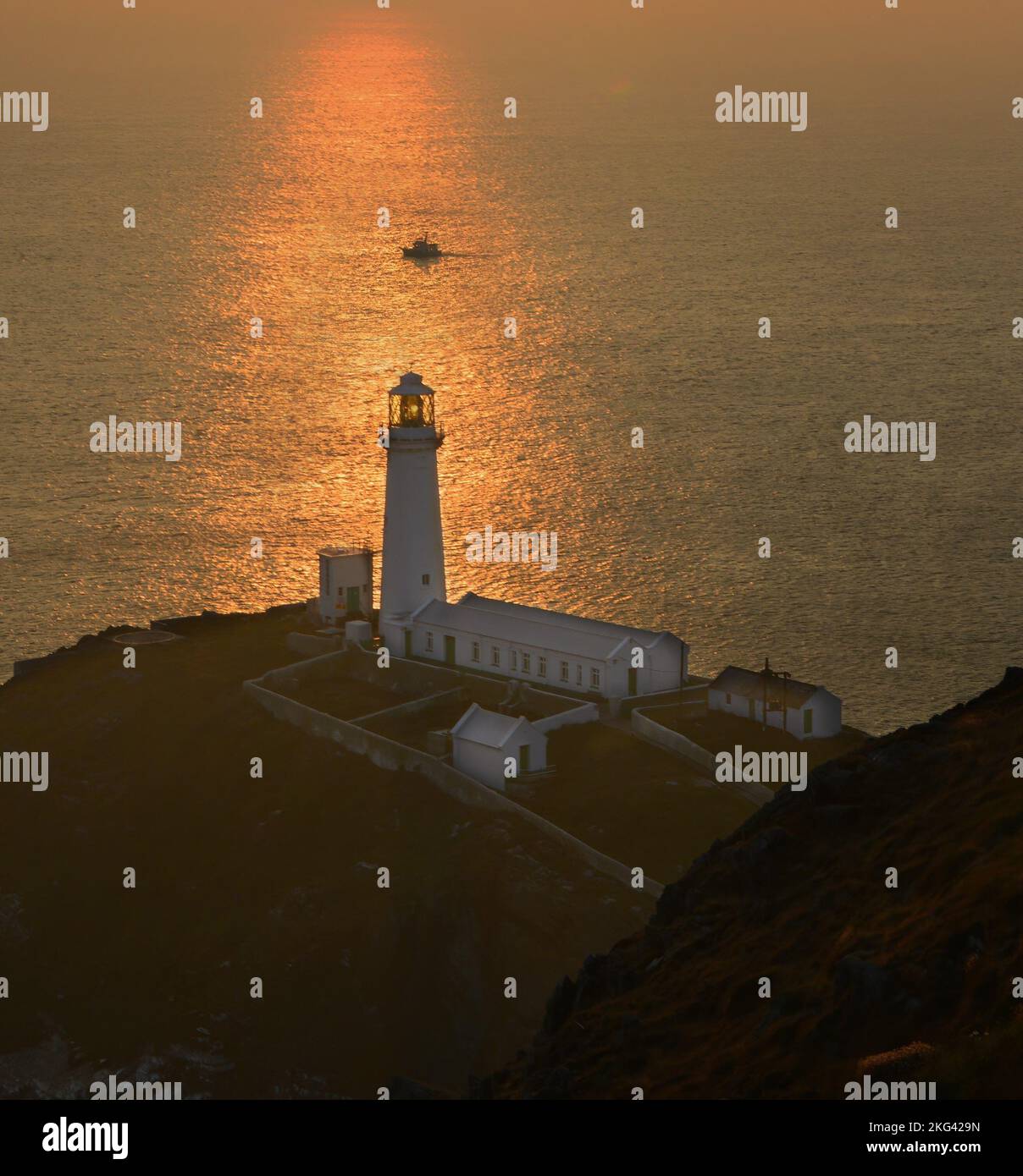 South Stack Lighthouse, Anglsey, North Wales, Großbritannien bei Sonnenuntergang an einem warmen Abend. Das Sonnenlicht bedeckt den Leuchtturm und ein vorbeifahrendes Boot. Stockfoto